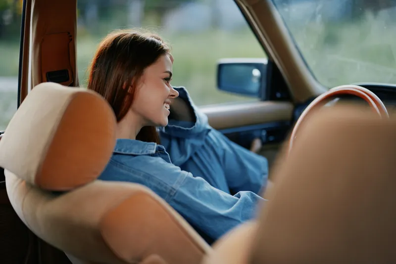 Smiling young woman in a cozy car interior wearing a denim shirt, conveying happiness and relaxation in a summer setting.