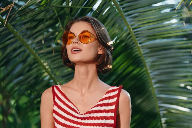 A stylish woman wearing a striped red and white top and large sunglasses poses outdoors among tropical greenery. She exudes confidence and joy, creating a bright, carefree summer mood.