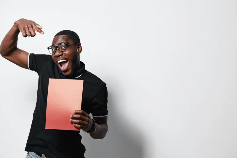 African American man with dark skin and glasses excitedly pointing at blank paper in hand, wearing black polo shirt against white background with joyful expression.