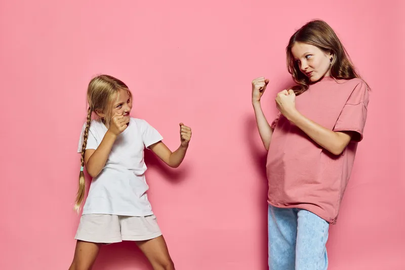 Two young girls playfully pose with fists raised against a pink background. Both show confident and playful expressions in casual clothing. Concept of childhood fun.