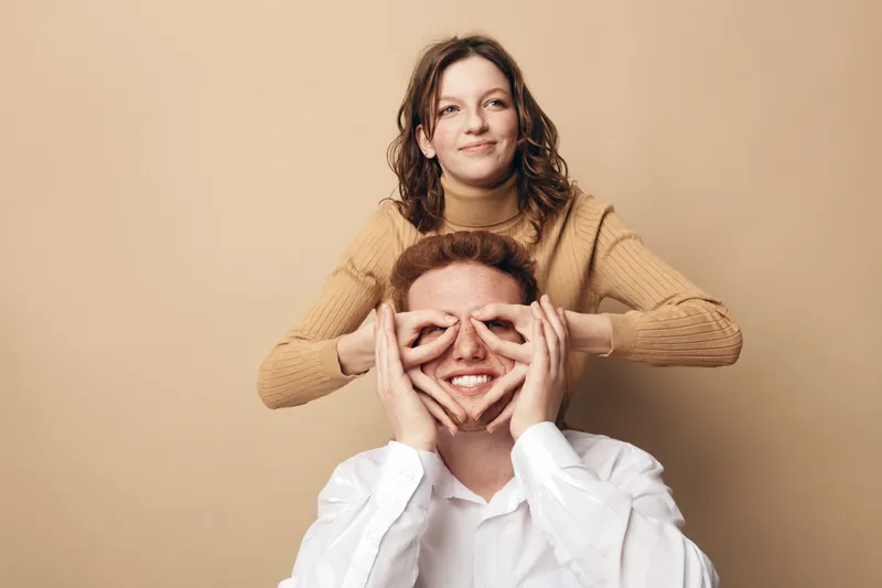 Young woman in brown sweater playfully covers eyes of smiling man in white shirt on beige background. Concept of friendship, fun, happiness, and casual lifestyle in studio setting.