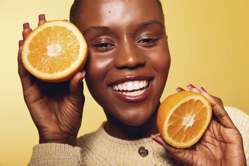 Happy African woman with dark skin holds two orange halves near face smiling brightly on yellow background. Concept of healthy lifestyle, fresh fruit, and natural beauty.