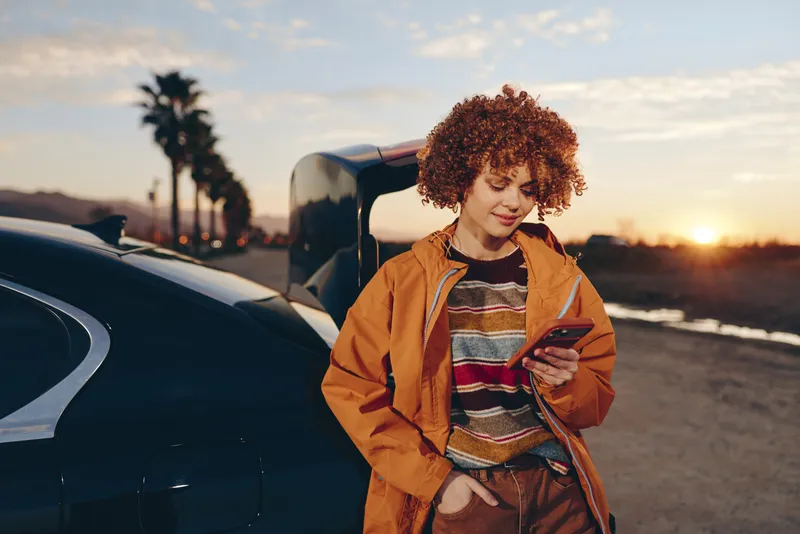 Woman lifestyle smartphone car sunset roadside street travel casual holding, curly haired woman stands beside parked car, looks at phone, warm golden hour light, outdoor road scenery at dusk