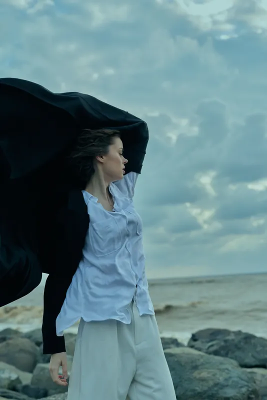 Woman on a rocky coast with a flowing cape in the wind, dramatic seaside scene, fashion movement and freedom against a cloudy ocean sky, atmospheric and expressive.