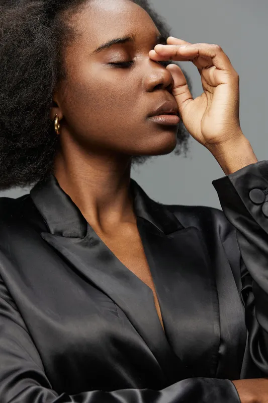 A young Black woman in a sleek black satin suit expressing calmness and introspection, with her eyes closed against a muted gray background