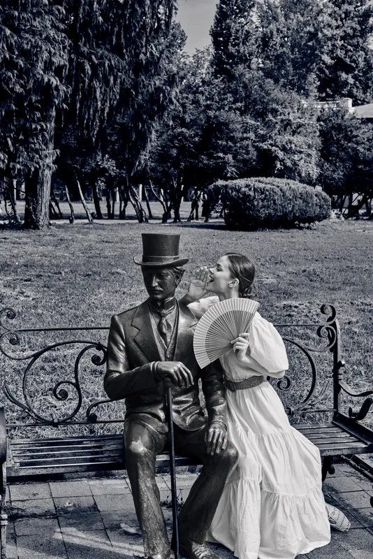A vintage statue of a man in a top hat sits on a park bench beside a woman in a flowing dress, sharing a quiet romantic moment in a black and white garden scene.