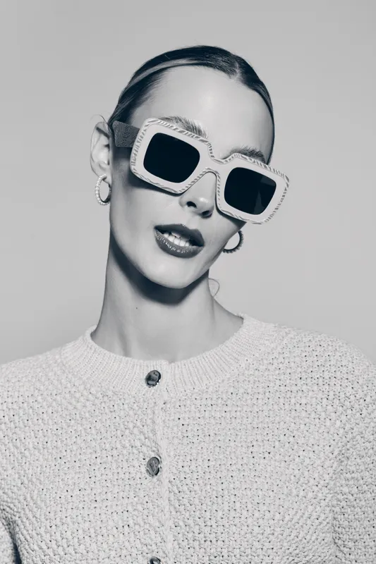 Stylish white woman with pale skin wearing large square sunglasses and knitted cardigan poses with head tilt in monochrome studio. Fashion portrait with modern eyewear concept.
