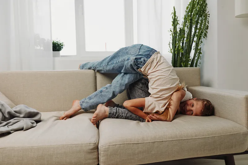 Two playful children in casual clothes having fun on beige sofa in bright living room with plants and natural light. Concept of childhood joy, family bonding, and indoor playtime.