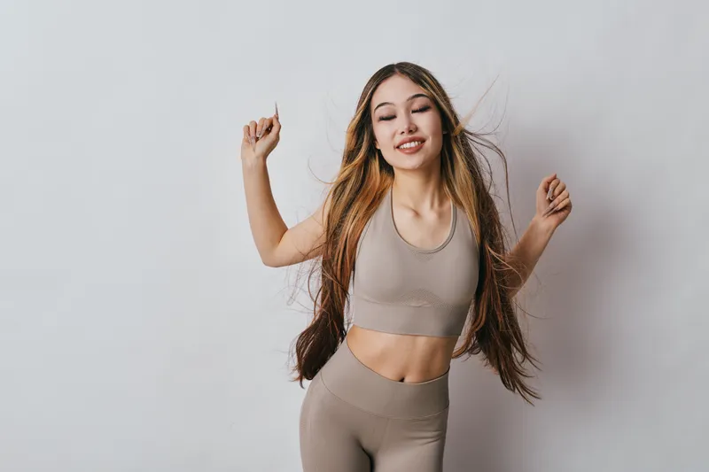 Young woman in sportswear dancing with eyes closed and smiling on light background. Healthy lifestyle, fitness, active, movement and joyful mood with long hair in studio.