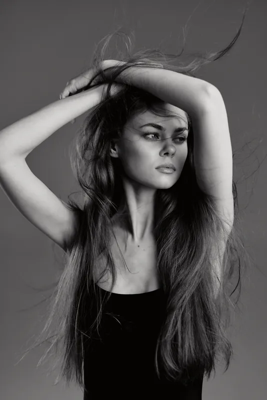 Black and white portrait of a serious young woman with long hair and arms raised, expressing elegance and intensity.