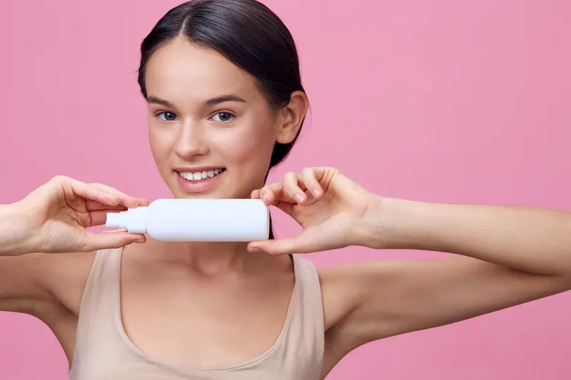 Smiling young woman holding a skincare bottle against a pink background, promoting healthy skincare and beauty concepts
