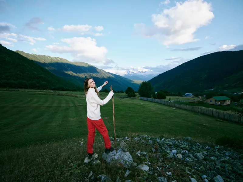 Hiking along a rocky trail in wide mountains and green countryside, a woman in white top and red pants explores outdoors, landscape, and nature during a bright adventure