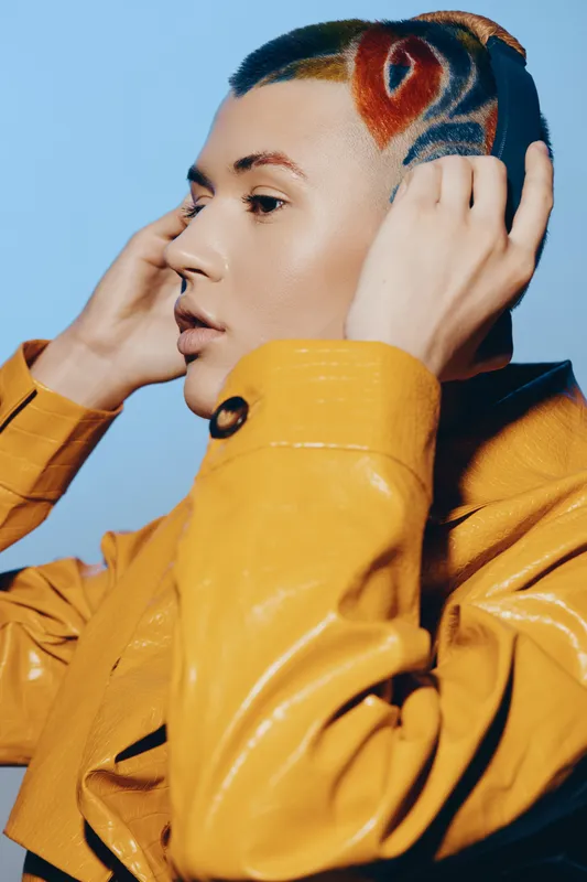 creative woman with colorful shaved hairstyle wearing yellow jacket touching headphones, profile portrait with blue background, modern fashion and unique look