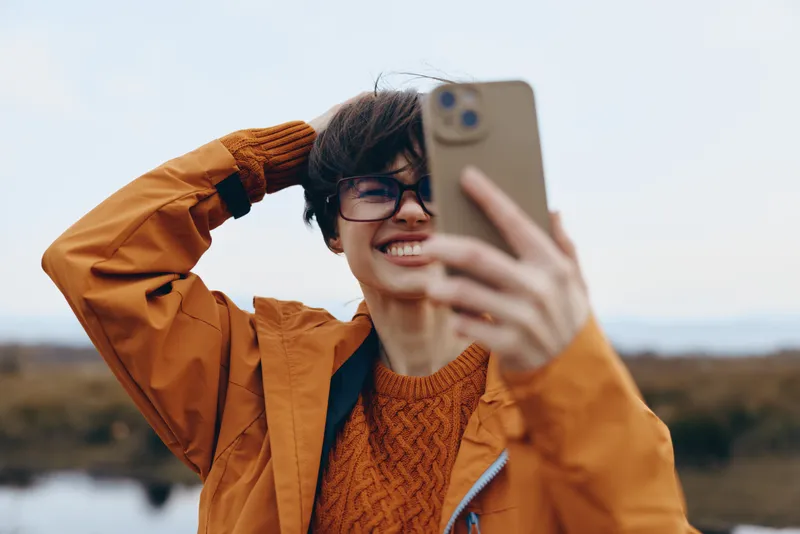 Woman lifestyle selfie outdoors, smiling in glasses, holding phone toward face for casual portrait, wearing orange jacket and knit sweater, bright daylight, distant hills in background