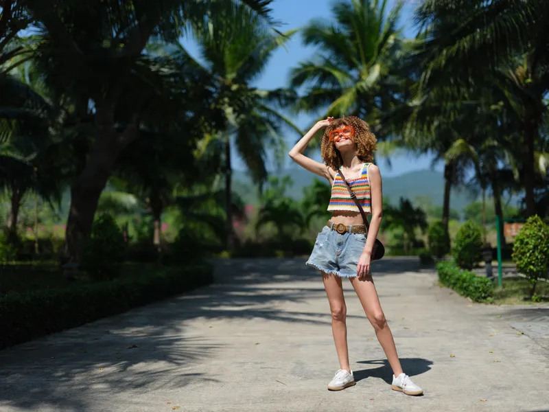 young woman with curly hair wearing colorful striped top and denim shorts standing on sunny pathway, shielding eyes from bright sunlight, tropical palm trees around