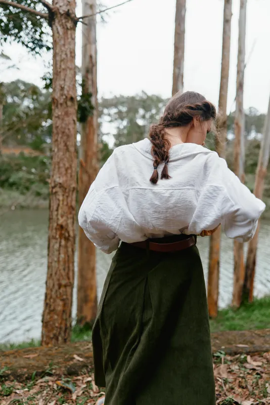 A woman in a white blouse and green skirt standing by a river surrounded by trees, expressing tranquility in a serene natural setting