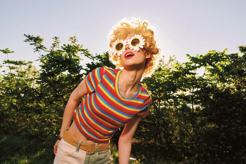 Vibrant outdoor portrait of a woman in a striped top and flower sunglasses, celebrating a carefree lifestyle with retro film color, sunny summer energy, and a playful candid pose.