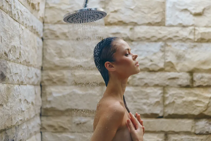 young woman enjoying outdoor shower with stone wall background, natural skin, wet hair, relaxation, serene expression, summer wellness concept