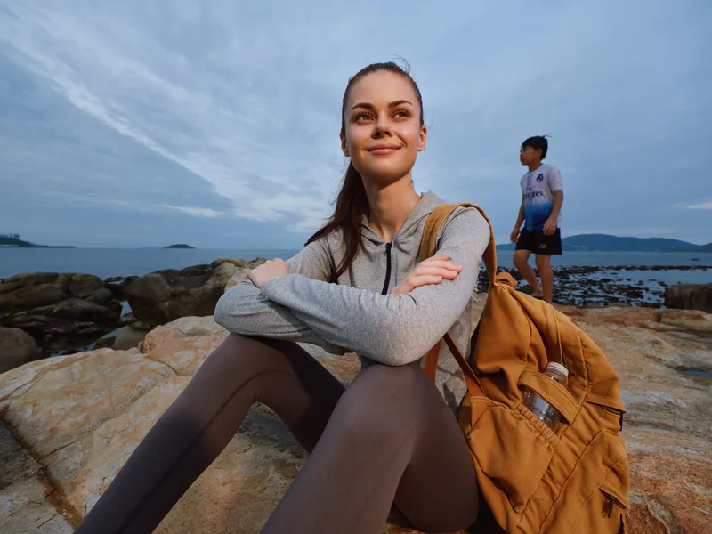 Young woman smiling on a rocky beach at sunset, wearing a gray hoodie and brown backpack, with a boy in the background, evoking a sense of adventure and tranquility