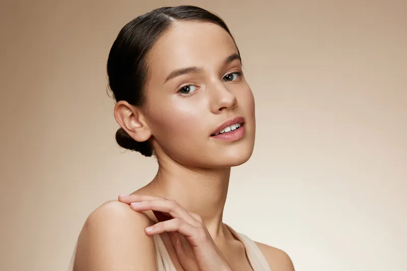 Elegant young woman with natural makeup poses gracefully against a neutral beige background, showcasing beauty and confidence with a soft smile and a minimalist style