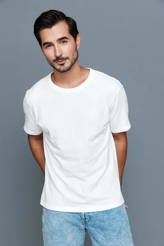 Stylish, smiling man in white t-shirt and blue jeans poses confidently against a solid gray background, showcasing a casual and modern look. People lifestyle concept.