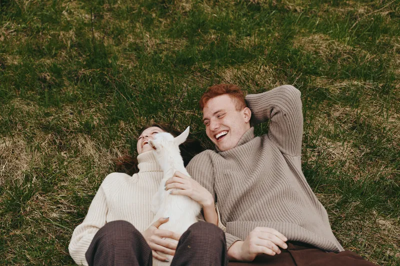Young couple lying on grass laughs joyfully while holding a small white baby goat, enjoying a relaxed lifestyle moment in cozy sweaters outdoors during autumn.