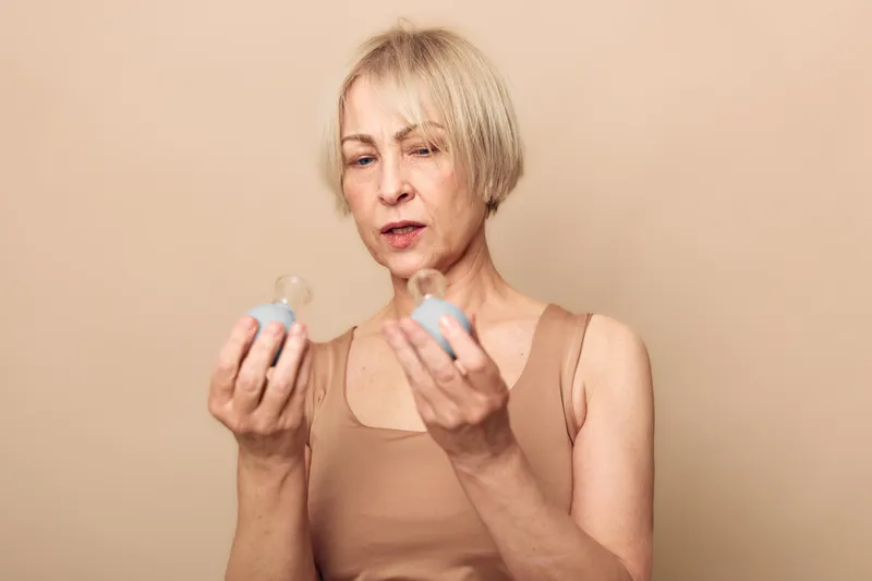Mature woman with short blonde hair examines two small bottles in a beige studio. Concept for decision making, skincare products, and personal care routine for middle aged adults.