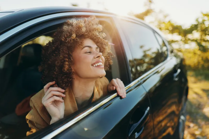 Young woman with curly hair smiles out a car window, basking in golden sunlight with retro film color, embodying carefree urban freedom and a playful, candid mood.