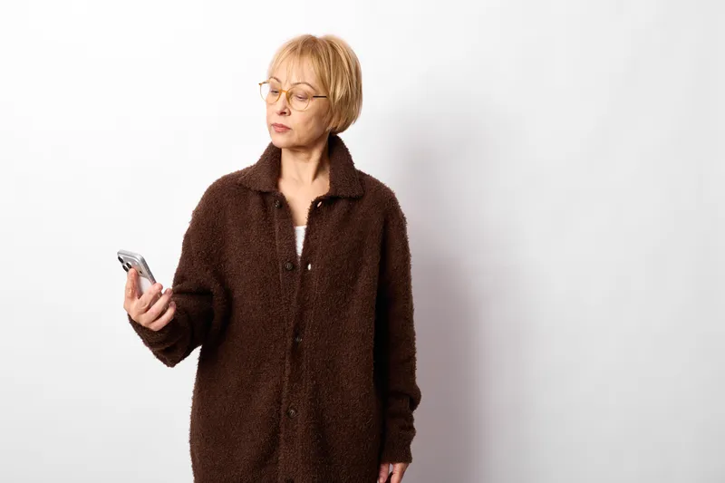 Elderly woman uses smartphone indoors, standing on clean white background, brown coat, short hair, thoughtful expression, simple portrait framing, soft studio light, calm pose