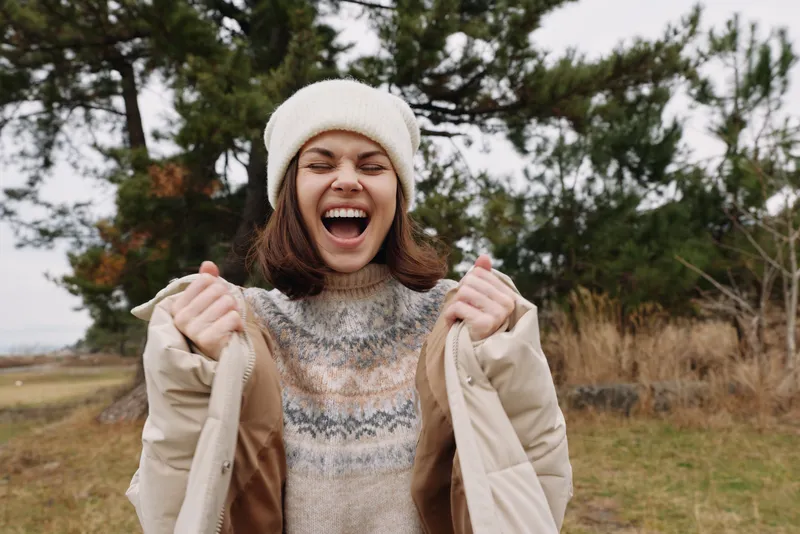 Joyful woman outdoors in a knit sweater and warm coat, laughing with hands in sleeves, autumnal field and trees in the background, a vibrant candid moment