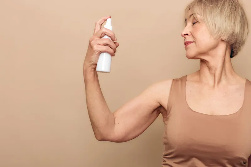 Mature woman in beige tank top sprays mist on her arm against neutral background. Concept of skincare, hydration, self-care, and healthy aging with natural beauty and wellness focus.
