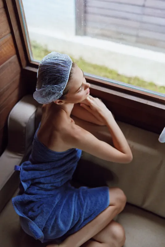 Relaxed woman with towel and shower cap sitting by window inside wooden sauna, enjoying peaceful moment with natural light and cozy atmosphere.