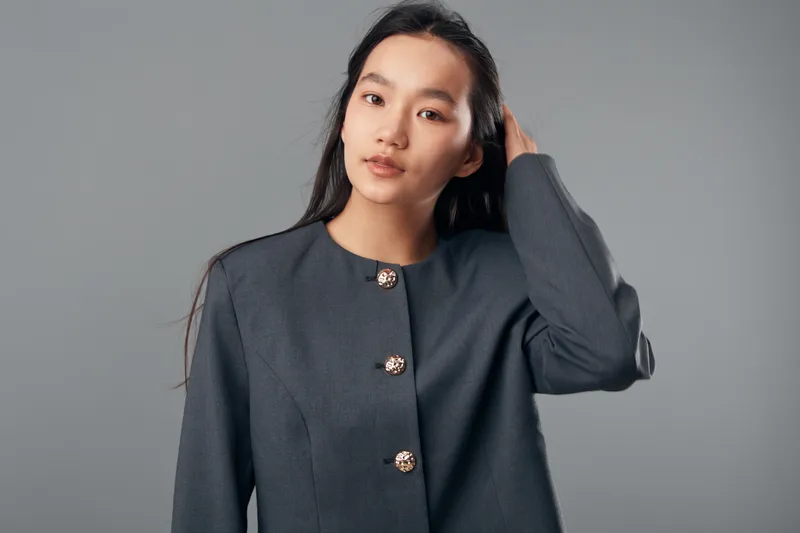 Portrait of young woman with long dark hair in stylish gray jacket posing with hand in hair against plain background. Model gazes at camera with calm confident expression in studio.