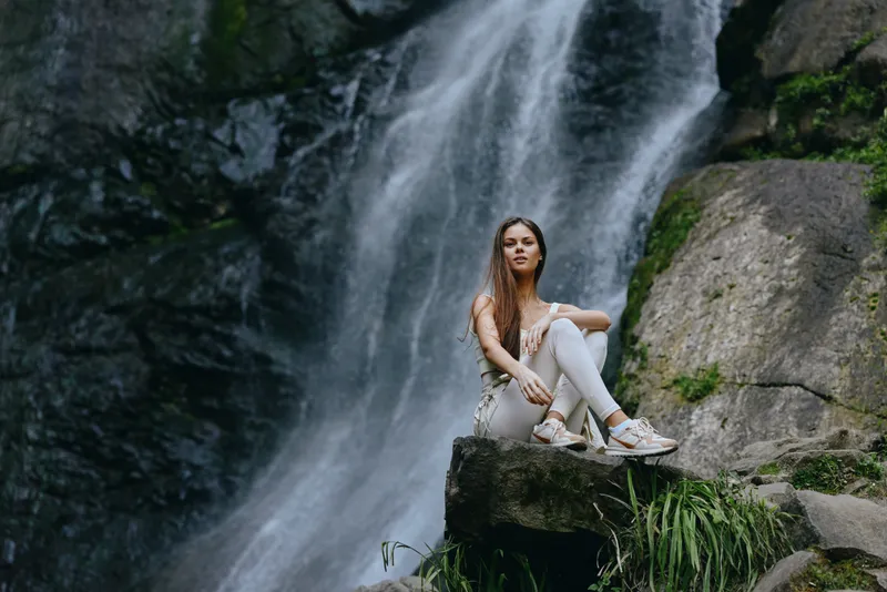 young woman sitting on rock near cascading waterfall surrounded by lush green plants and natural stone landscape in outdoor setting