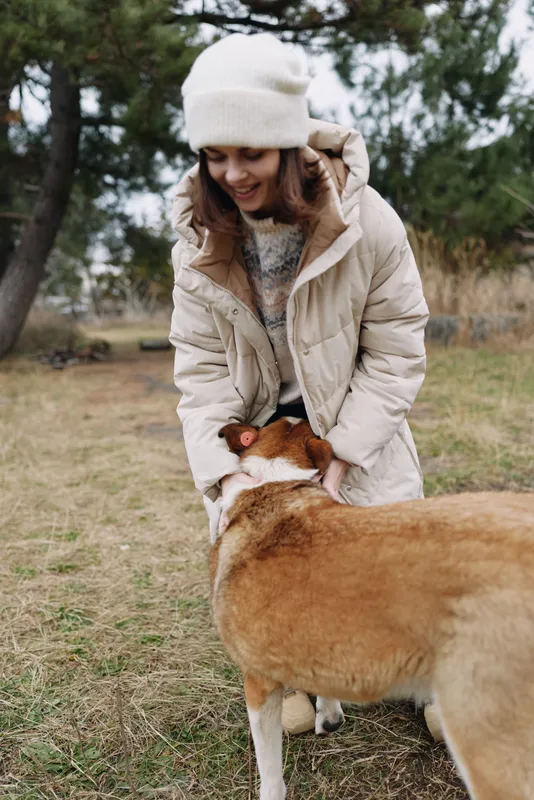 Woman in a warm coat and beanie crouches to greet a large dog, enjoying a playful moment outdoors in a park on a chilly day.