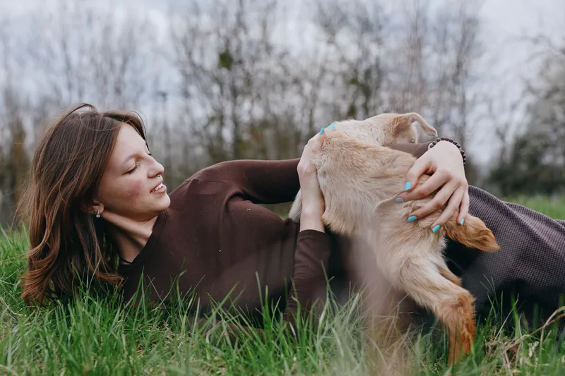 A woman lies in grass softly embracing a goat kid outdoors, capturing affection and tenderness in a natural setting, highlighting nature, lifestyle, and mutual care between human and animal