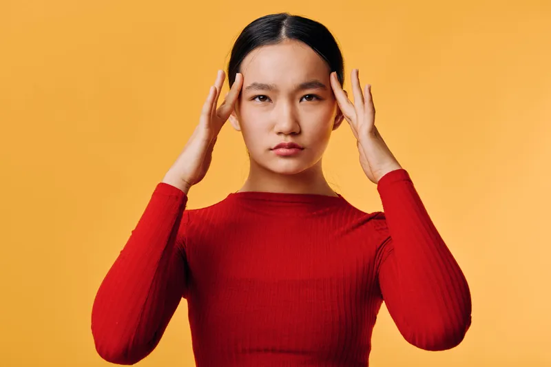 Woman indoors in red long sleeve shirt touching her temples with serious expression and focused look on orange background for stress or headache concept.