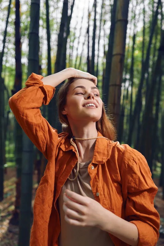 Woman portrait in bamboo forest smiling with eyes closed, orange jacket and casual clothing, nature outdoors scene showing peaceful joy, relaxed model enjoying sunlight and serenity