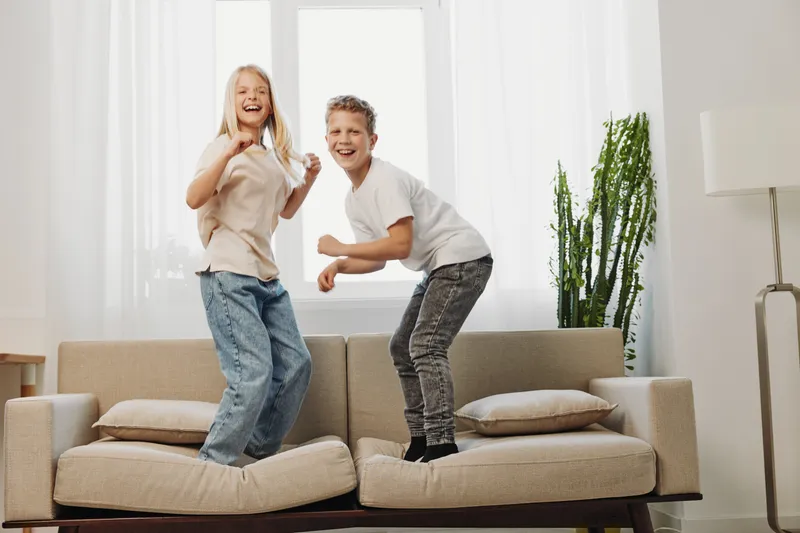 Happy children jump on couch in bright living room, laughing and having fun. Concept for childhood joy, playful moments, and carefree indoor activity at home.