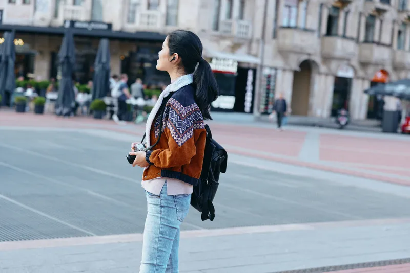 Woman with backpack on a city street captures a casual urban moment, fashion inspired outfit, standing with a camera ready, exploring an outdoor plaza and architecture