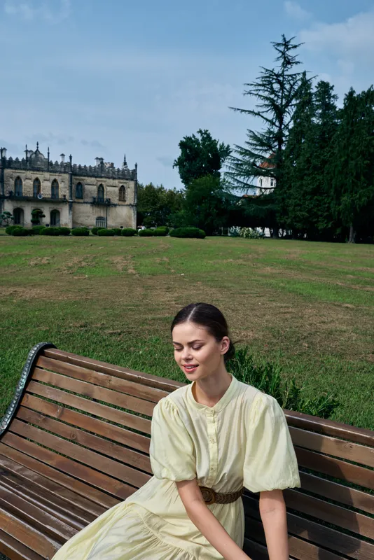 Woman in a vintage pale dress sits on a wooden bench in a park with a historic castle in the background, creating a calm outdoor portrait with timeless charm