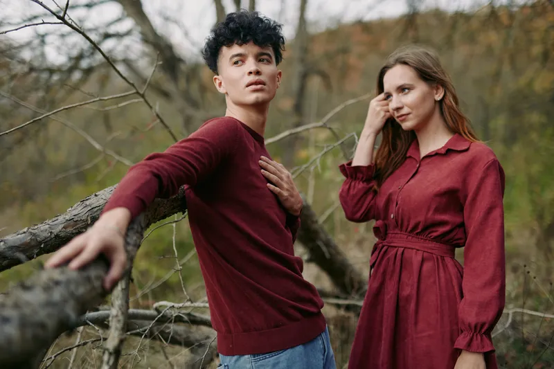 Young couple in matching burgundy outfits pose naturally outdoors, conveying genuine connection and relaxed presence, focused on real emotions and honest interaction in a rustic woodland setting.