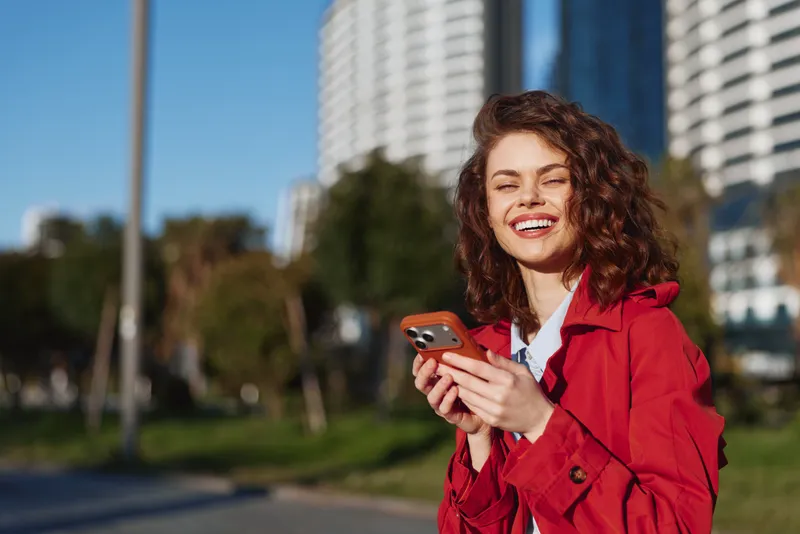 A smiling woman in a red coat uses a smartphone outdoors in an urban park, bright day, reflecting casual urban life and connection.
