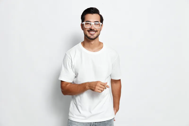 Confident smiling young man wearing glasses and a white t-shirt, pointing at the camera, isolated on plain solid background. Modern lifestyle concept