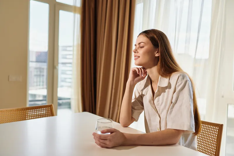 Woman portrait at home, sunlight on table as she sits holding a glass in relaxation during morning. Cozy interior with curtains, thoughtful peaceful mood and natural bright light.
