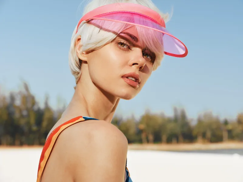 Portrait of a stylish young woman outdoors wearing a pink visor, sunlit skin, and a colorful strap. Clean composition highlights confidence, summer vibe, and carefree mood.