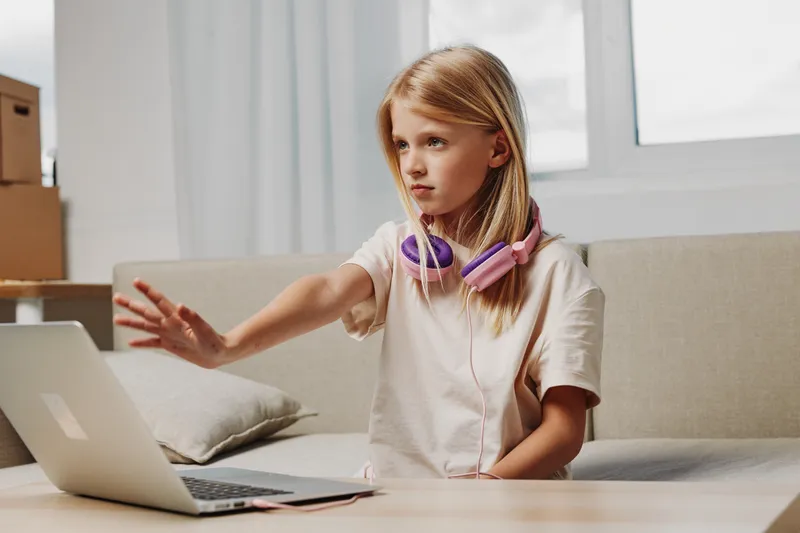 Blond girl with headphones sitting on sofa uses laptop with hand raised in stop gesture in cozy bright room. Concept of technology, childhood, communication, and online interaction.