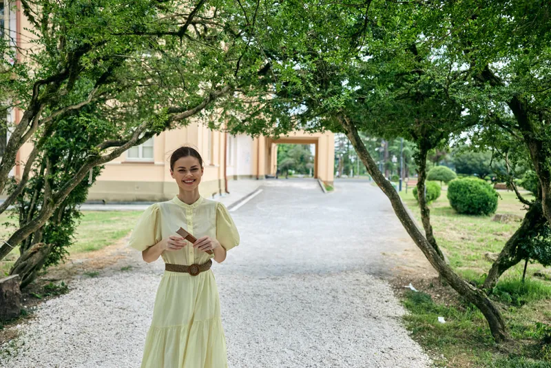woman dress garden walkway portrait authenticity inclusivity, smiling woman in pale vintage dress stands under green arching trees on gravel path