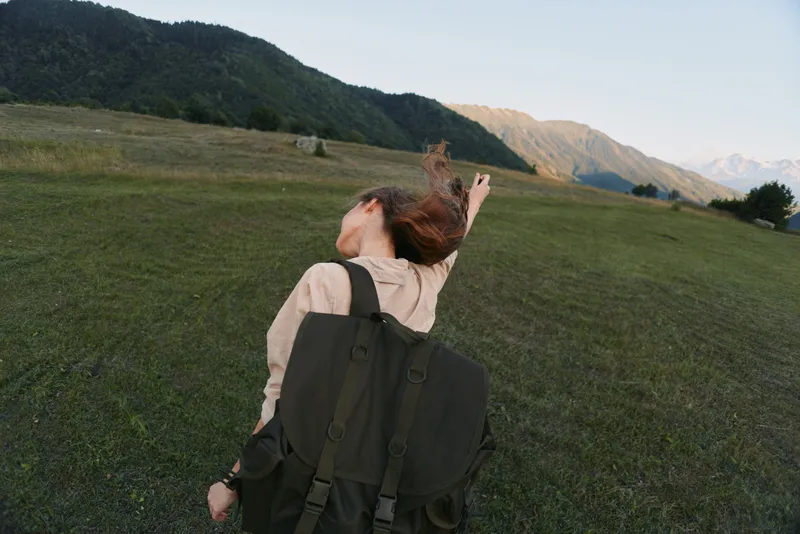 Joyful woman outdoors, backpack on shoulder, arms lifted, sunlit meadow with distant mountains, carefree lifestyle, natural colors, gentle breeze, exploring moment today
