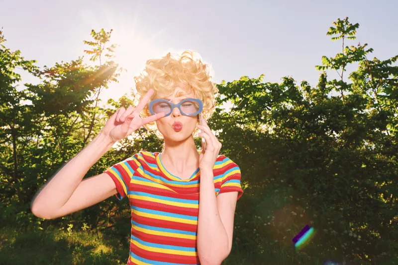 Woman outdoors wearing a striped shirt and retro sunglasses, sunlit moment with laughter, vibrant colors against greenery, casual lifestyle with nostalgic film color